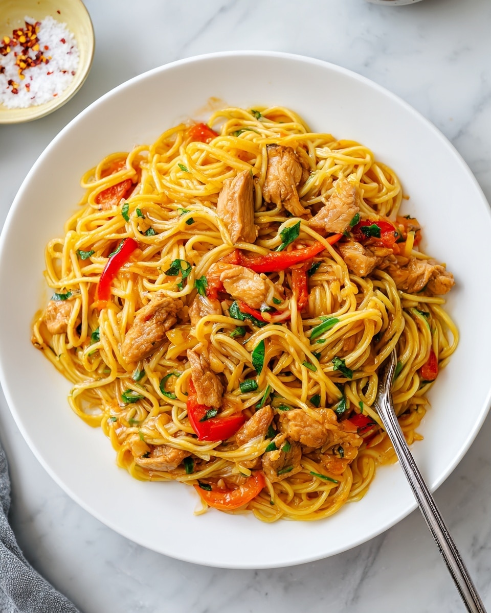 A white plate is filled with a nest of yellow pasta strands mixed with pieces of light brown cooked chicken and thin slices of bright red bell pepper, all coated in a shiny sauce. Small bits of green herbs are scattered throughout the dish. A silver fork rests on the right edge of the pasta, slightly inserted into the noodles. The plate sits on a white marbled surface. Nearby, a small round white plate with more pasta and a pale yellow dish containing coarse salt with red flakes are partially visible. photo taken with an iphone --ar 4:5 --v 7