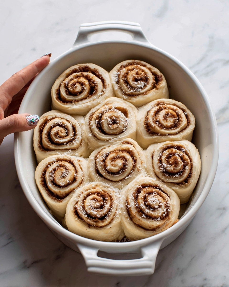 A white oval baking dish filled with eleven unbaked cinnamon rolls arranged close together, each roll showing visible spiral layers of light dough and brown cinnamon filling swirling from the center outward. The rolls have a slightly rough texture with some white flour dust on the surface. A woman's hand with decorated nails holds the bottom handle of the dish. The scene is set on a white marbled surface. photo taken with an iphone --ar 4:5 --v 7