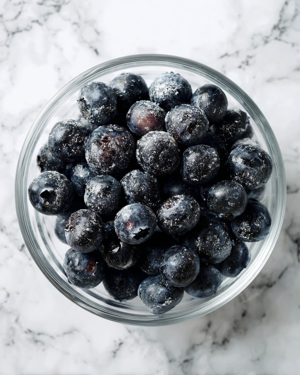 A clear glass bowl filled with one layer of dark blue-black blueberries that are dusted with a light white powder, placed on a white marbled surface. Photo taken with an iphone --ar 4:5 --v 7