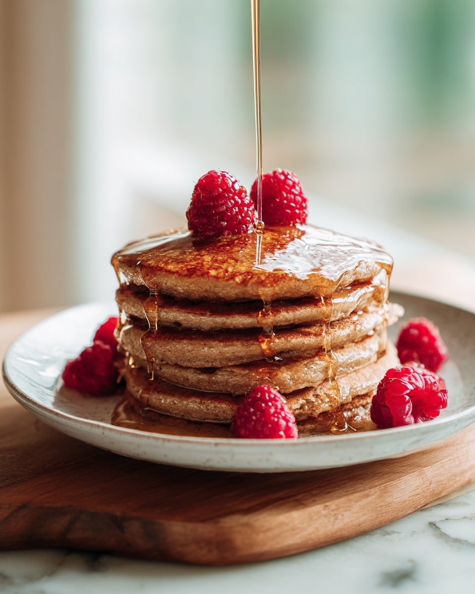 A stack of four brown pancakes is placed in the center of a white plate with scalloped edges. On the top pancake, there are three whole red raspberries that add a bright pop of color. A silver fork on the right side of the plate holds a bite-sized piece of pancake with one raspberry on top, and a silver knife with a white handle lies next to it. Light golden syrup is drizzled over the pancakes and pools around the base on the plate. The plate is set on a white marbled surface, and a light pink cloth napkin is visible on the right edge of the image. Photo taken with an iphone --ar 4:5 --v 7