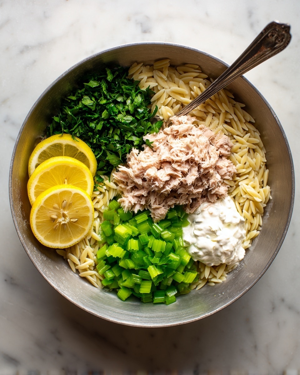 A large metal bowl holds a mix of light beige orzo pasta as the base layer. On top of the pasta, there are bright green chopped celery pieces scattered in the center. Next to the celery, on the right side, is a mound of light brown tuna. Below the tuna, there is a dollop of white creamy spread, likely mayonnaise or yogurt. To the left of the celery, there are fresh dark green chopped herbs, and slices of yellow lemon with visible seeds sit on the pasta at the top and bottom left edges. A metal spoon is resting inside the bowl with its handle extending out. The whole scene is set on a white marbled surface. Photo taken with an iphone --ar 4:5 --v 7