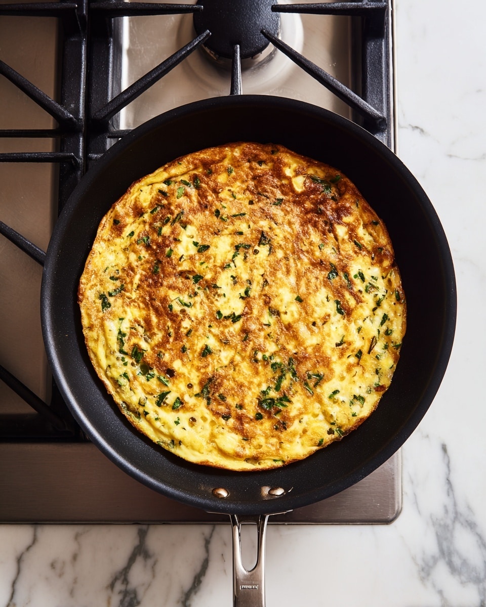 A black frying pan sits on a gas stove with a cooked frittata inside, filling the pan in one even layer. The frittata is golden yellow with small green herbs spread throughout, and the top shows a slightly browned texture with some crispy edges. The pan handle extends out to the right side, and the stove’s metal grates are visible under and around the pan. The background is a white marbled surface. photo taken with an iphone --ar 4:5 --v 7