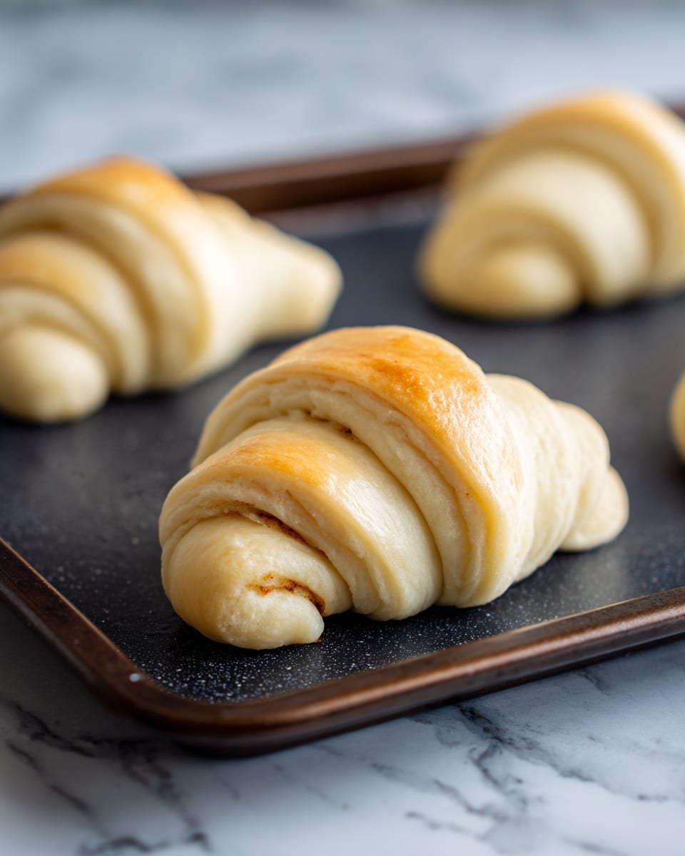 A close-up view of an unbaked crescent roll on a dark baking tray, showing one roll in the center with smooth, pale dough wrapped in a layered spiral around a cinnamon and sugar filling, with the edge tucked in neatly; two more similar rolls are blurred in the background on the same baking tray. The surface beneath the tray is a white marbled texture. Photo taken with an iphone --ar 4:5 --v 7