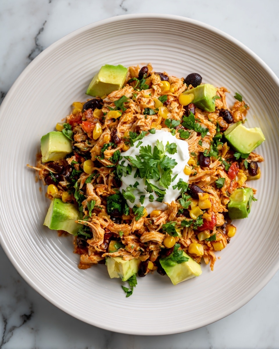 A white ridged plate holds a mound of shredded chicken mixed with black beans, yellow corn, and small pieces of red bell pepper, creating a mix of orange, black, yellow, and red colors. Green chopped scallions and cilantro are scattered throughout and on top. Small cubes of bright green avocado are placed around the edges and on the chicken mix. A dollop of creamy white sauce sits in the middle of the dish. The plate rests on a white marbled surface. photo taken with an iphone --ar 4:5 --v 7