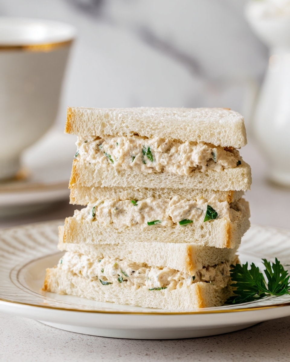 Three triangular sandwich layers are stacked on a white plate with a simple patterned rim, each layer made of soft white bread with a light beige filling that looks creamy and slightly textured, possibly tuna or chicken salad, with hints of green herbs inside. The background has a white marbled texture, and behind the plate, there is a blurry white cup with a gold rim. The overall look is soft and fresh. photo taken with an iphone --ar 4:5 --v 7