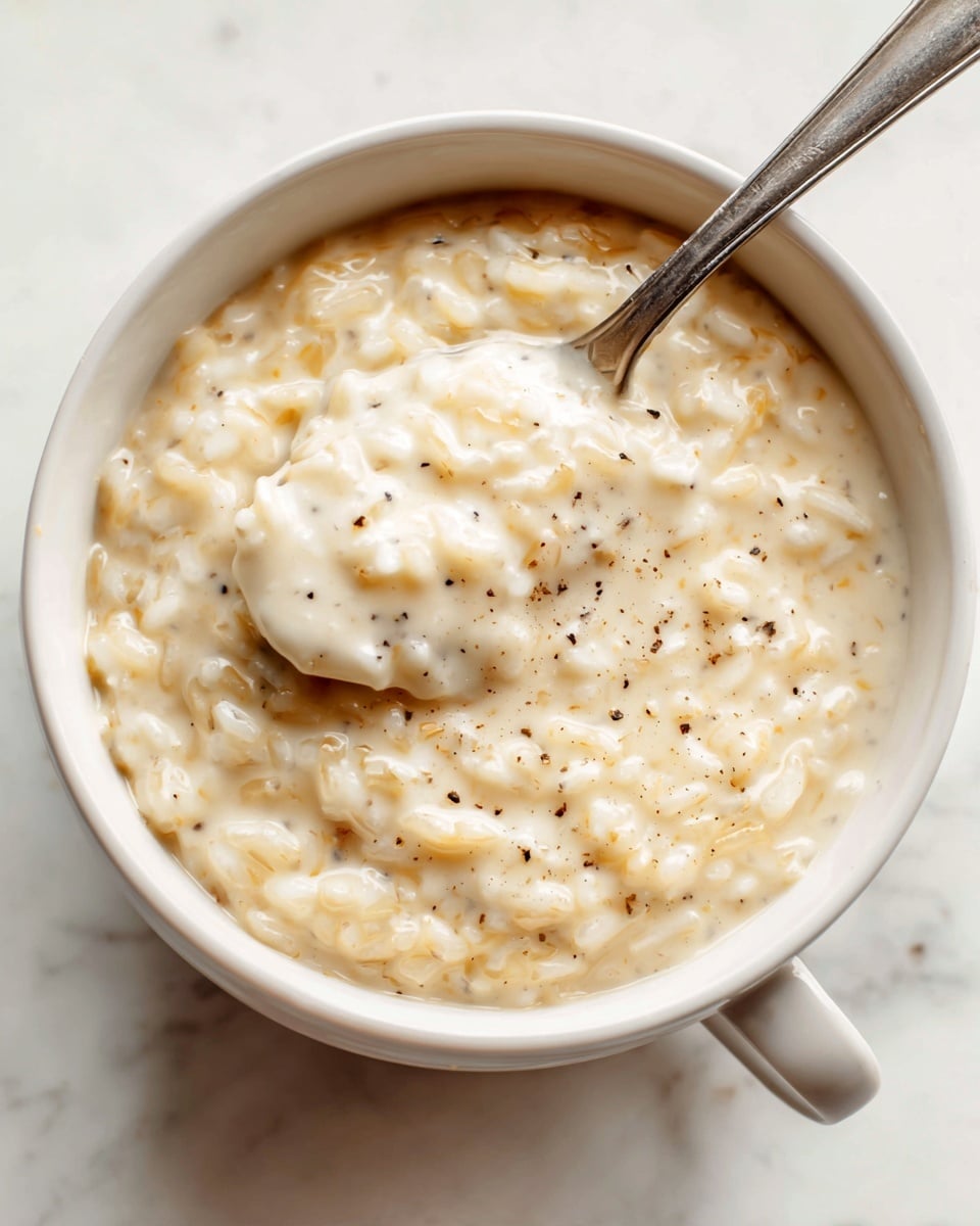 A close-up of a white bowl filled with creamy rice pudding that is thick and slightly lumpy, showing soft, tender grains of rice mixed with a smooth, light beige custard-like sauce. The pudding has tiny black vanilla specks scattered throughout, adding texture and subtle color contrast. A metal spoon rests inside the bowl, scooping up a portion of the pudding, with some pudding clinging to the sides of the bowl. The bowl sits on a white marbled surface, enhancing the warm and comforting feel of the dish. photo taken with an iphone --ar 4:5 --v 7