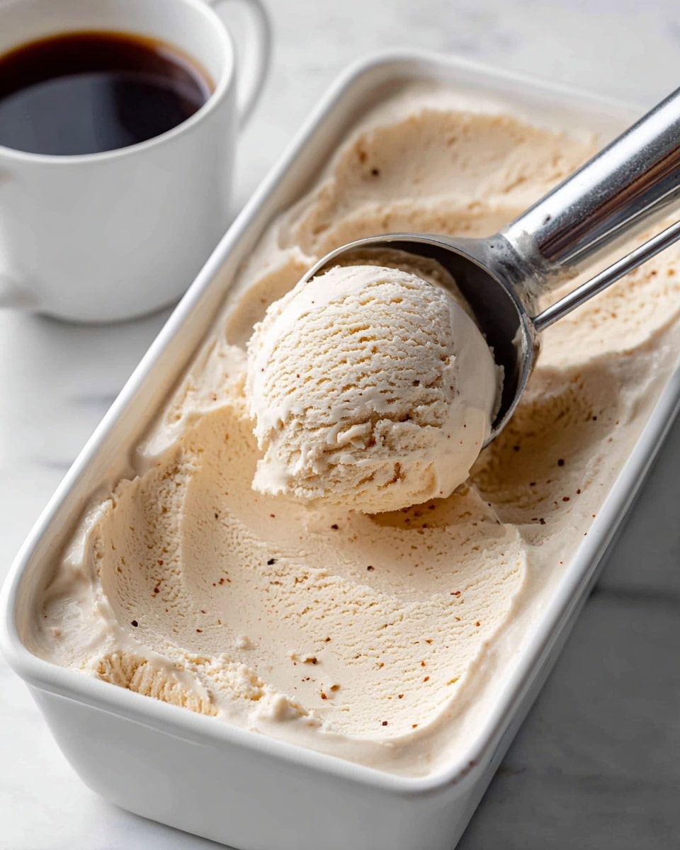 A close-up of a white rectangular dish filled with light brown ice cream that has a smooth and creamy texture with some small dark specks visible throughout. A metal ice cream scooper lifts a single round scoop showing soft, slightly rough edges on the ice cream. The dish is placed on a white marbled surface, and a small white cup holding dark liquid is partially seen on the left side. photo taken with an iphone --ar 4:5 --v 7