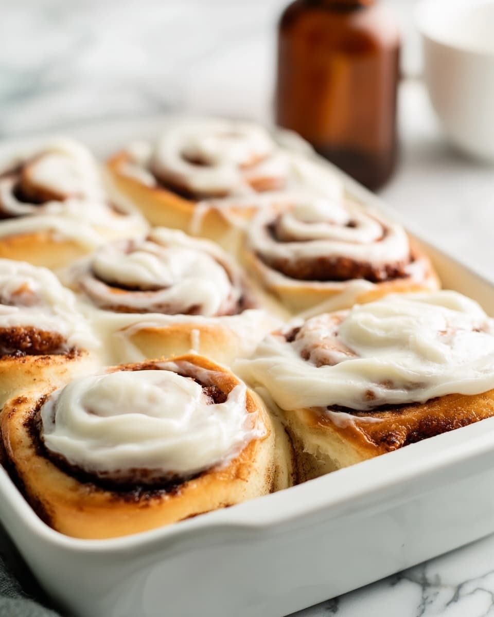 A close-up of a cinnamon roll with three visible layers showing soft, light brown dough swirled with dark cinnamon filling. The top is covered with thick, creamy white icing spread unevenly, revealing the cinnamon spiral underneath. The roll sits on a white plate with a textured surface, all placed on a white marbled texture. Photo taken with an iphone --ar 4:5 --v 7