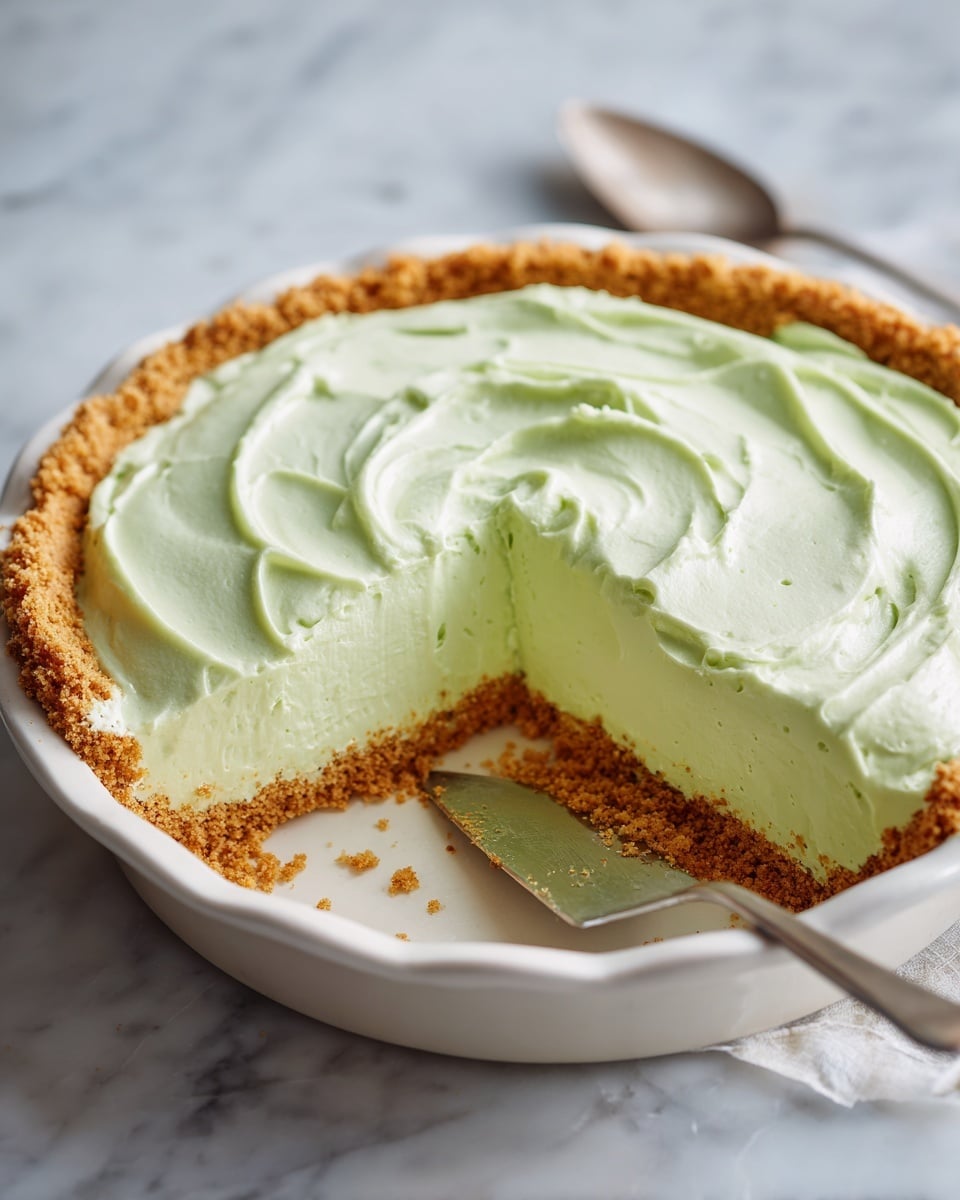 A pie with three layers is shown, placed on a white marbled surface. The bottom layer is a crumbly golden brown crust that lines the pie dish, holding the filling in place. The middle layer is a thick, pale green creamy filling with a smooth texture, and the top layer is the same pale green cream spread evenly with soft swirls and slight peaks. One slice has been removed, revealing the thickness and firmness of the filling compared to the crust. The pie dish is white and round, showing a clear contrast between the layers. Photo taken with an iphone --ar 4:5 --v 7