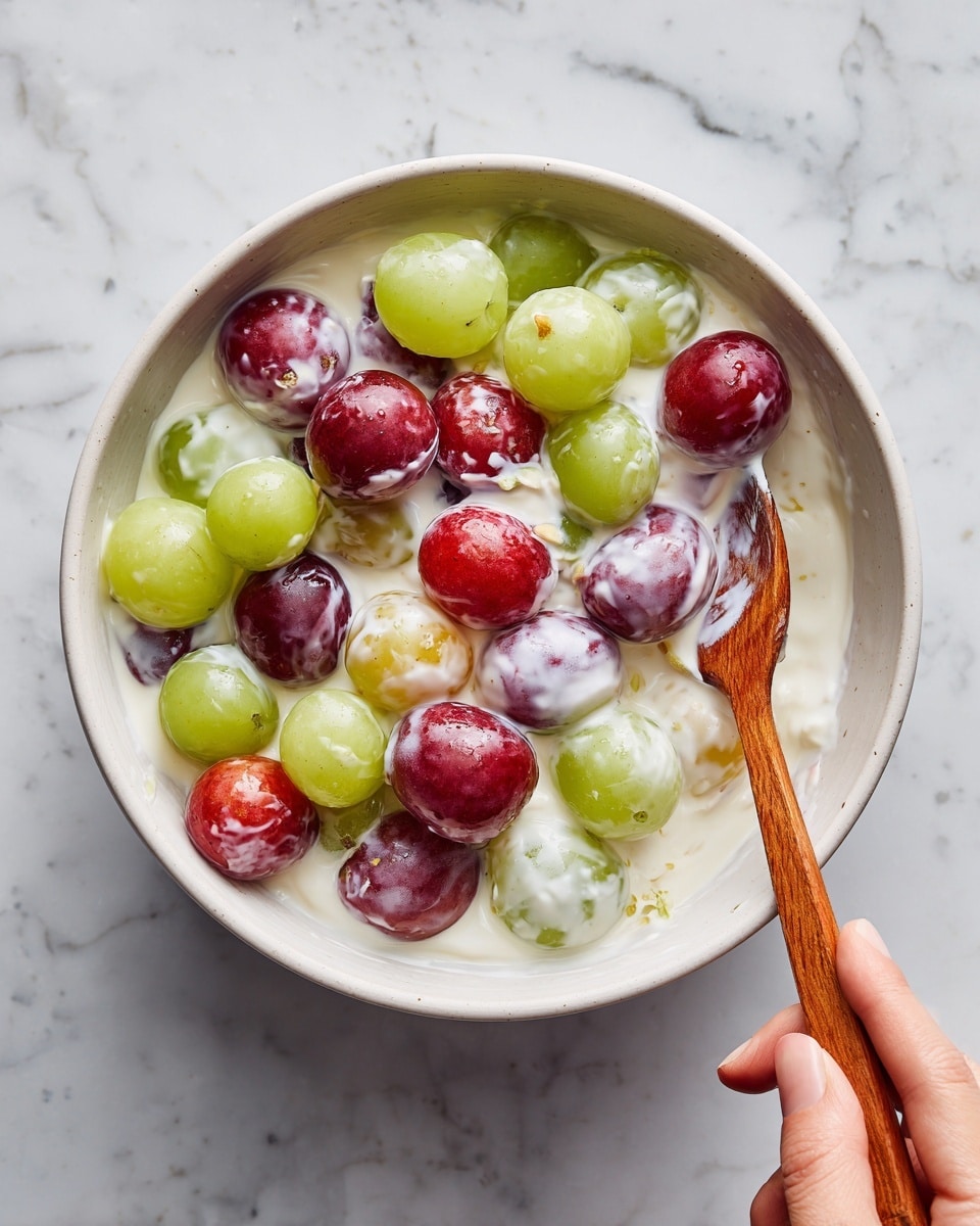 A white bowl filled with a creamy mixture covered in white yogurt-like sauce. Inside the sauce are whole grapes of two colors: green and red, with some red grapes showing their inner pale color where sliced. A wooden spoon is partially immersed in the mixture, lifting some of it. A woman's hand holds the spoon, stirring the grapes and sauce together. The bowl is placed on a white marbled surface background. photo taken with an iphone --ar 4:5 --v 7