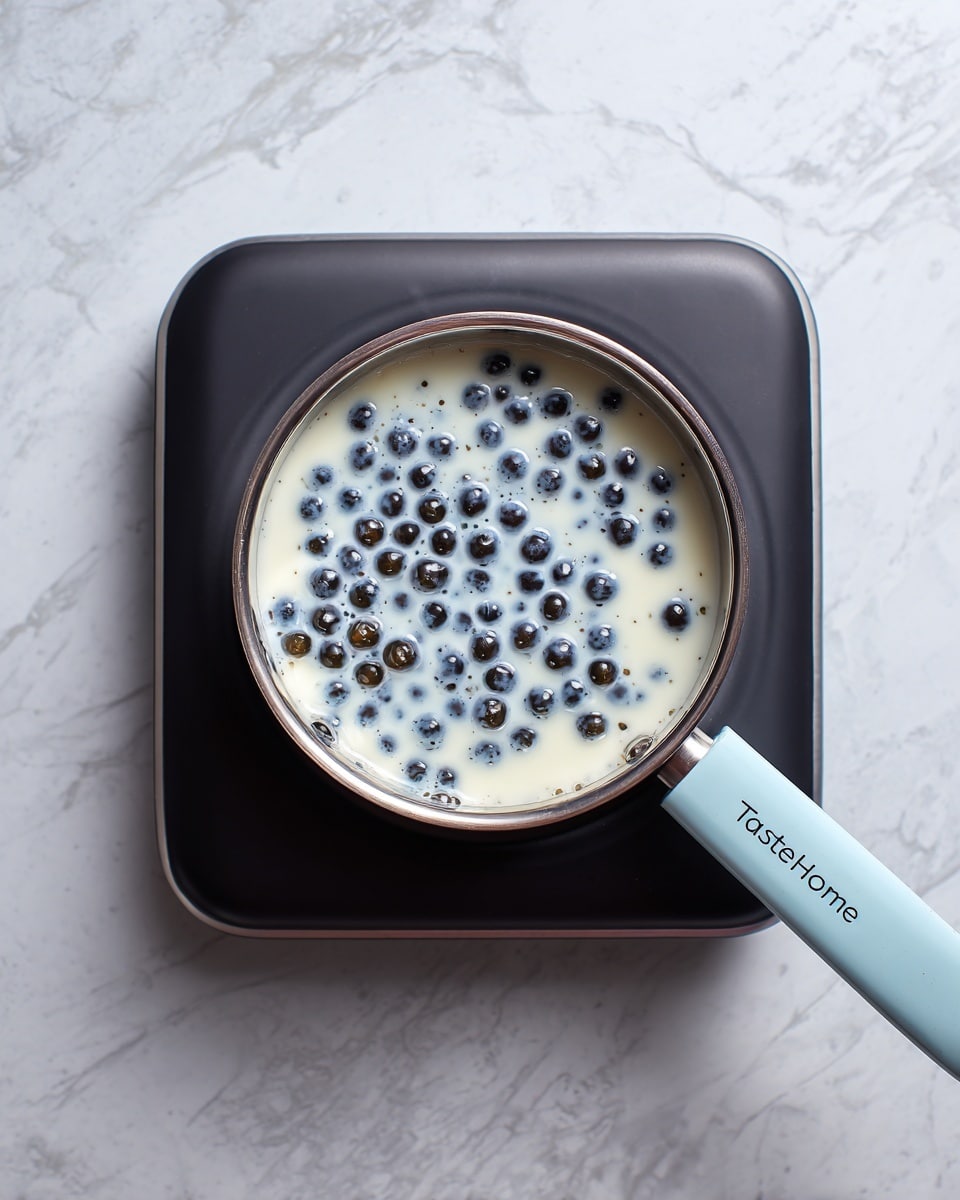 A close-up top view of a small silver saucepan sitting on a black hot plate, filled with a liquid that is mostly white with some cloudy spots and many small, round, shiny black pearls floating on top. The saucepan handle is light blue with