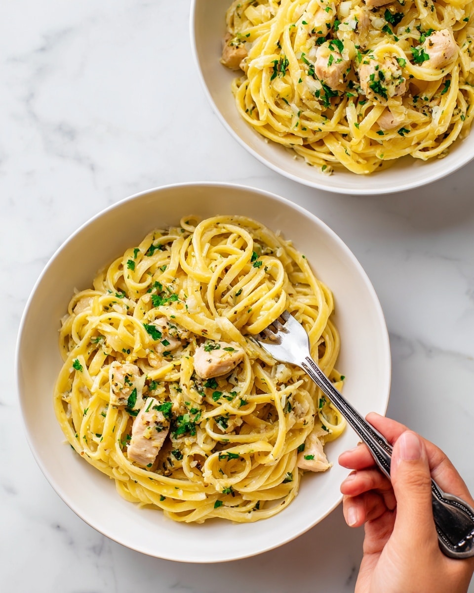 The image shows two white bowls filled with thick yellow pasta noodles arranged in loose piles, each topped with two pieces of light brown cooked chicken sprinkled with green herbs and visible small bits of garlic or onion. One bowl is fully visible, with a fork held by a woman's hand scooping a small amount of pasta from the right side, while the other bowl is partially shown on the left edge. The background is a white marbled surface, creating a clean and bright setting. photo taken with an iphone --ar 4:5 --v 7