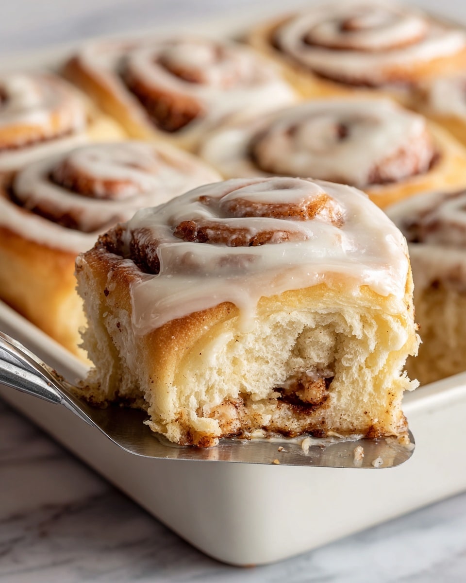 The image shows a close-up of a cinnamon roll being lifted by a metal spatula above a white tray full of other cinnamon rolls. The cinnamon roll has three visible layers: the outer layer is creamy white icing with a smooth texture, the middle layer is a soft golden brown dough, and the inner swirl has a darker brown cinnamon filling. The background is a white marbled surface, softly blurred to keep focus on the cinnamon roll. photo taken with an iphone --ar 4:5 --v 7