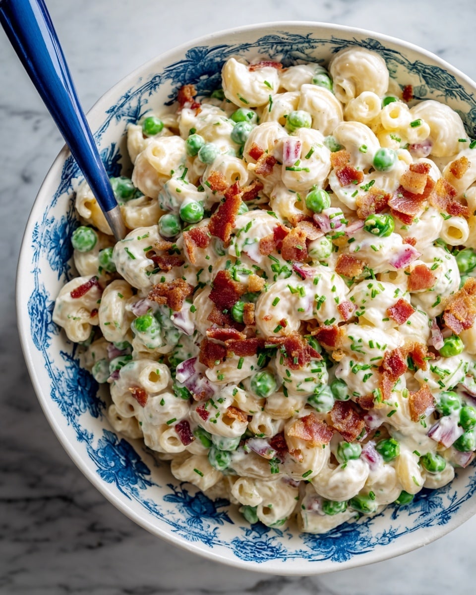 The image shows a close-up of a creamy macaroni salad in a white bowl with blue floral patterns. The salad has three main layers: curved macaroni pasta coated in a thick white mayonnaise sauce, bright green peas scattered throughout, and small pieces of crispy browned bacon on top. There are also some chunks of pale yellow cheese and bits of red onion mixed in. A blue spoon is partially submerged in the salad on the left side. The bowl is placed on a white marbled surface with a lace cloth partially visible in the background. Photo taken with an iphone --ar 4:5 --v 7