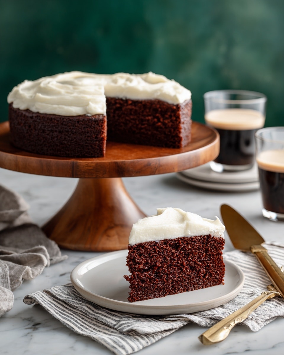 A single slice of chocolate cake with one thick, smooth layer of creamy white icing on top sits on a white plate. The cake is dark brown and looks soft with a slightly rough texture. A gold fork lies next to the cake slice on the plate. The plate is on a striped cloth with a white marbled surface underneath. In the blurred background, there is another plate with a similar slice and a dark drink in a clear glass. Photo taken with an iphone --ar 4:5 --v 7