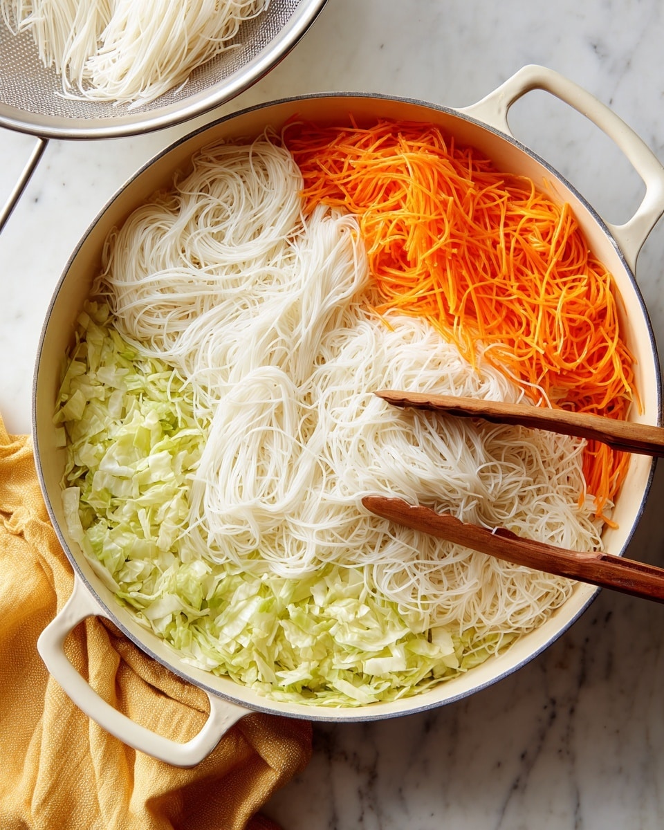 A round white pan is on a white marbled surface, filled with three visible layers of food. The bottom layer is chopped light green cabbage, spread evenly across the pan. On top of this is a layer of thin, shredded orange carrots scattered evenly. The top layer consists of long, white rice noodles loosely placed over the vegetables. Two wooden kitchen tongs rest in the pan on the right side, slightly pressing into the white noodles. To the left of the pan, a metal colander holding more white rice noodles sits on a yellow cloth. The scene shows preparation before mixing, with cooking on an induction stove beneath the pan. Photo taken with an iphone --ar 4:5 --v 7