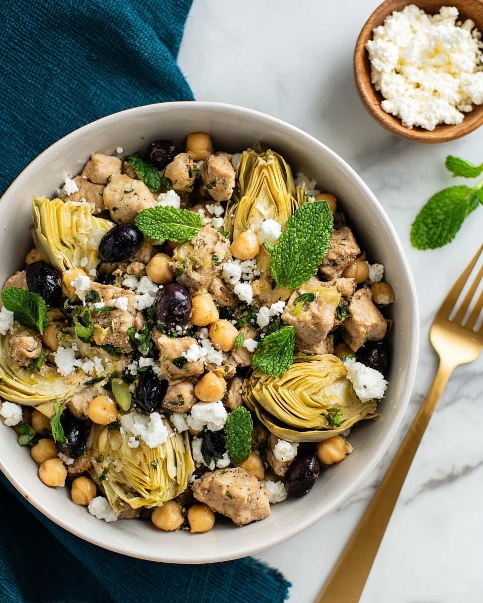 A white bowl filled with a colorful mix of cooked ingredients including browned meat chunks, beige chickpeas, and artichoke hearts with a soft yellow color and layered petal texture. The bowl also contains small dark olives and is topped with white crumbled cheese and bright green mint leaves scattered on top. The bowl sits on a dark teal cloth on a white marbled surface, next to a wooden fork and a small round white bowl filled with more crumbled white cheese. Photo taken with an iphone --ar 4:5 --v 7