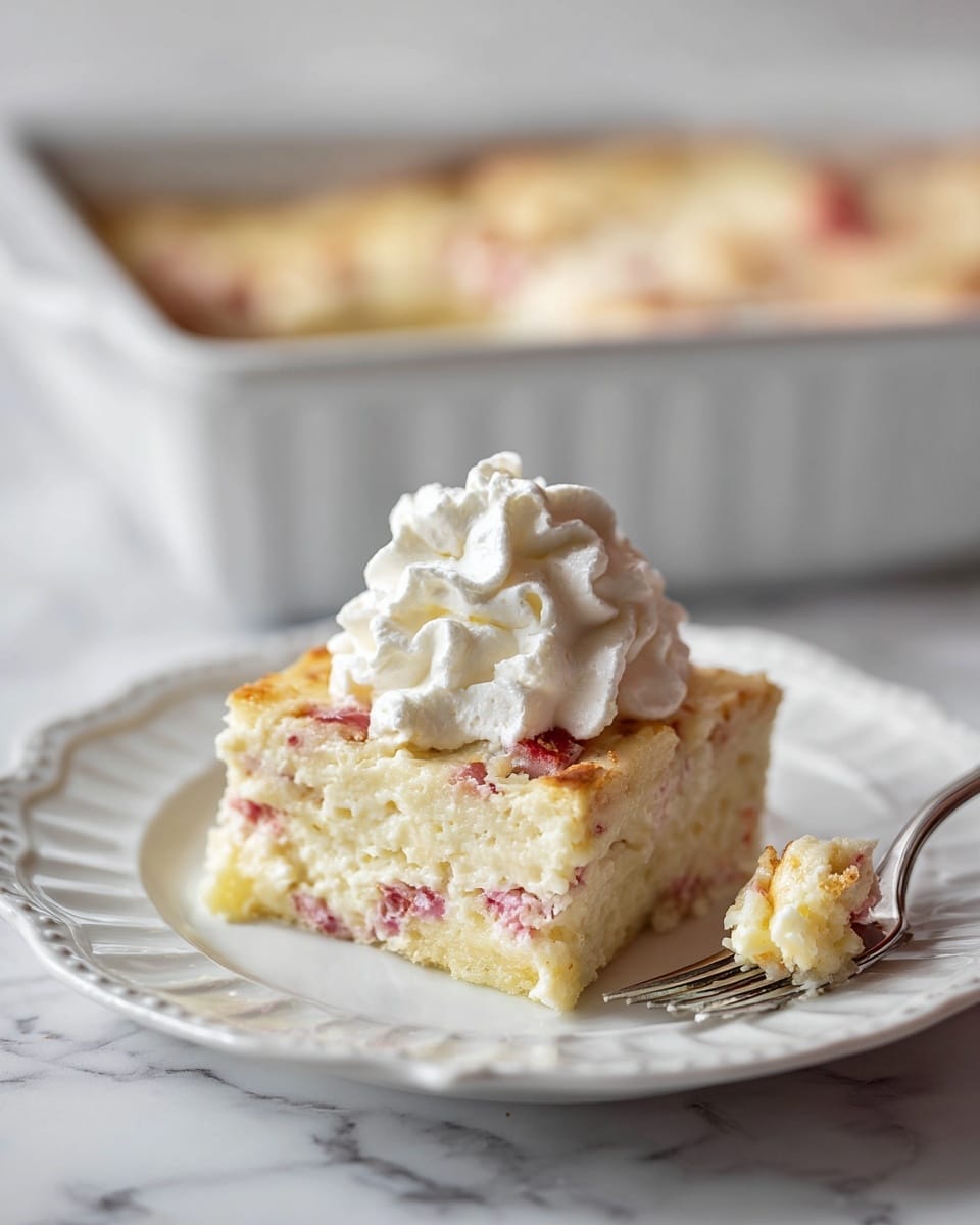 A close-up image of a square piece of creamy baked dish with visible small pink pieces inside, showing three layers: a golden-brown top layer, a middle layer with a creamy white color mixed with pink bits, and a slightly browned bottom layer. The dish is on a white plate with a scalloped edge. On top of the square piece is a big dollop of white whipped cream with a soft, fluffy texture. A silver fork holds a small bite of the dish with yellow and pink colors, placed on the right side of the plate. In the blurry background, there is a white baking dish filled with more of the same dish on a white marbled surface. photo taken with an iphone --ar 4:5 --v 7