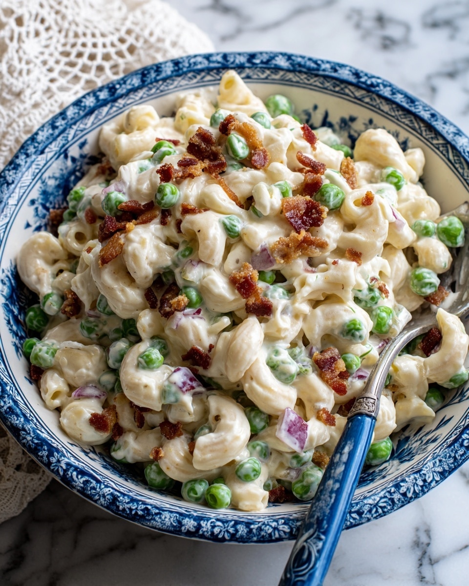 The image shows a close-up of a creamy macaroni salad in a white bowl with blue floral patterns. The salad has three main visible layers: curved elbow macaroni coated in a thick, white creamy dressing forms the base layer, mixed with bright green peas that add pops of color throughout. Scattered on top and mixed in are small, crispy-looking brown bacon bits and some pieces of purple-red onion, adding texture and contrast. The blue spoon rests inside the bowl, partly covered by the salad. The background is a white marbled surface with a bit of a lace cloth visible on the upper left corner. Photo taken with an iphone --ar 4:5 --v 7