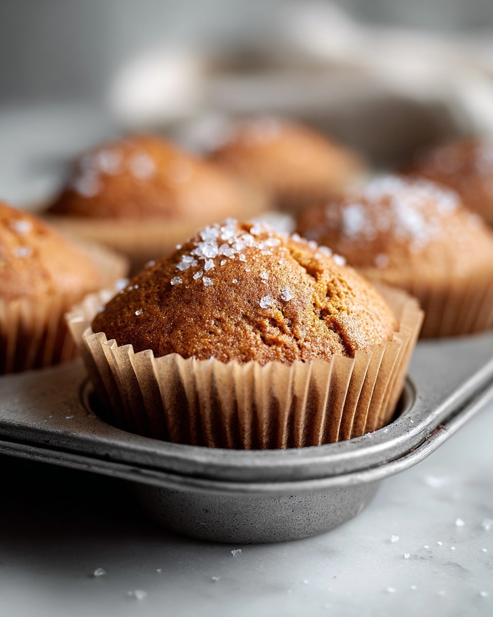 A close-up of a single brown muffin with a rough top sprinkled with sugar crystals, sitting in a light brown paper liner inside a grey metal muffin tray. Around it, other similar muffins are slightly out of focus, placed in the tray. The scene is set on a white marbled surface with a light, blurry background, showing a soft, natural light shining on the muffins. photo taken with an iphone --ar 4:5 --v 7
