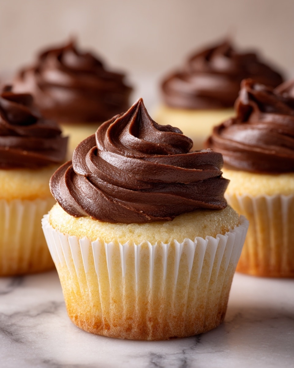 A cupcake with three layers: the base layer is light yellow cake with a soft texture, wrapped in a white paper liner; the middle layer is a thin dark brown layer on top of the cake; the top layer is thick, smooth, dark brown chocolate frosting, swirled in a spiral shape with pointed peaks. The cupcake is set on a white marbled surface with a plain light background. photo taken with an iphone --ar 4:5 --v 7