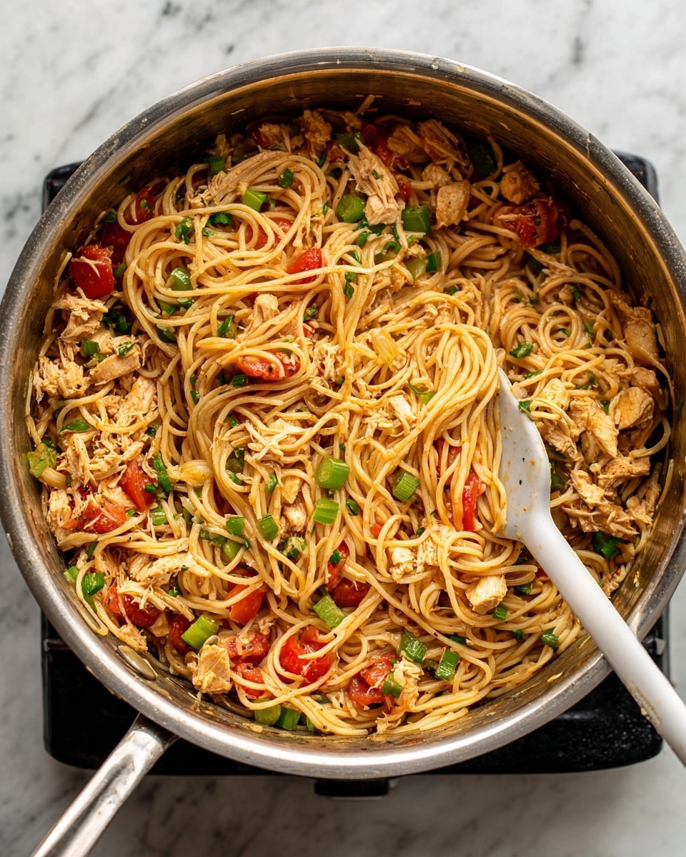 Inside a silver pot on a black stove burner, there is one layer of cooked spaghetti mixed with shredded light beige chicken, diced bright green celery, and chunks of red tomatoes. The texture of the food looks soft and moist, with the noodles swirling in the mix. A white spatula with a black handle is in the pot, lifting some of the spaghetti and vegetables. The scene is set on a white marbled surface that surrounds the pot. photo taken with an iphone --ar 4:5 --v 7