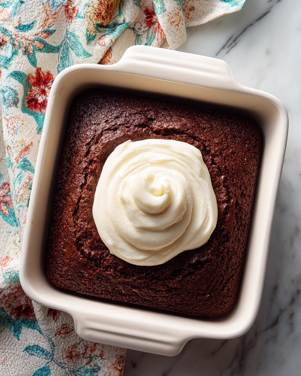 A single-layer dark brown chocolate cake is shown in a square white baking dish. The cake surface is cracked slightly, showing a dense texture. On top, near the center, there is a smooth round dollop of thick white frosting swirled gently. The dish rests on a white marbled surface with parts of a colorful cloth visible in the background. Photo taken with an iphone --ar 4:5 --v 7
