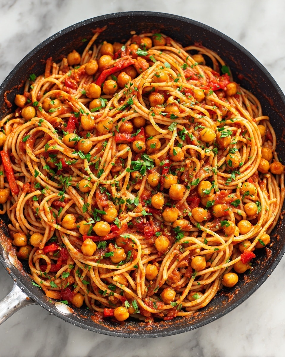 A close-up of a black pan filled with spaghetti cooked in a red tomato-based sauce mixed with round light beige chickpeas, small diced orange and red bell peppers, and green chopped herbs sprinkled on top. The spaghetti strands are coated evenly in the sauce, sitting in a glossy and thick orange-red liquid with visible lentils. The dish sits on a white marbled surface. photo taken with an iphone --ar 4:5 --v 7