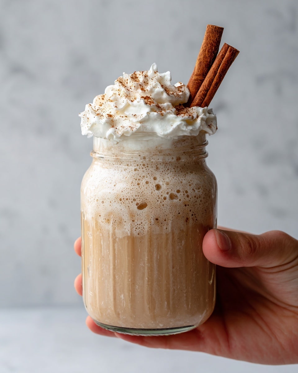 A creamy light brown drink fills a clear glass jar, with some foam bubbling at the top edge. On top of the drink is a thick, white whipped cream layer sprinkled with light brown powder, and two cinnamon sticks stand upright on one side. A woman's hand holds the jar from the bottom, against a white marbled textured background. photo taken with an iphone --ar 4:5 --v 7