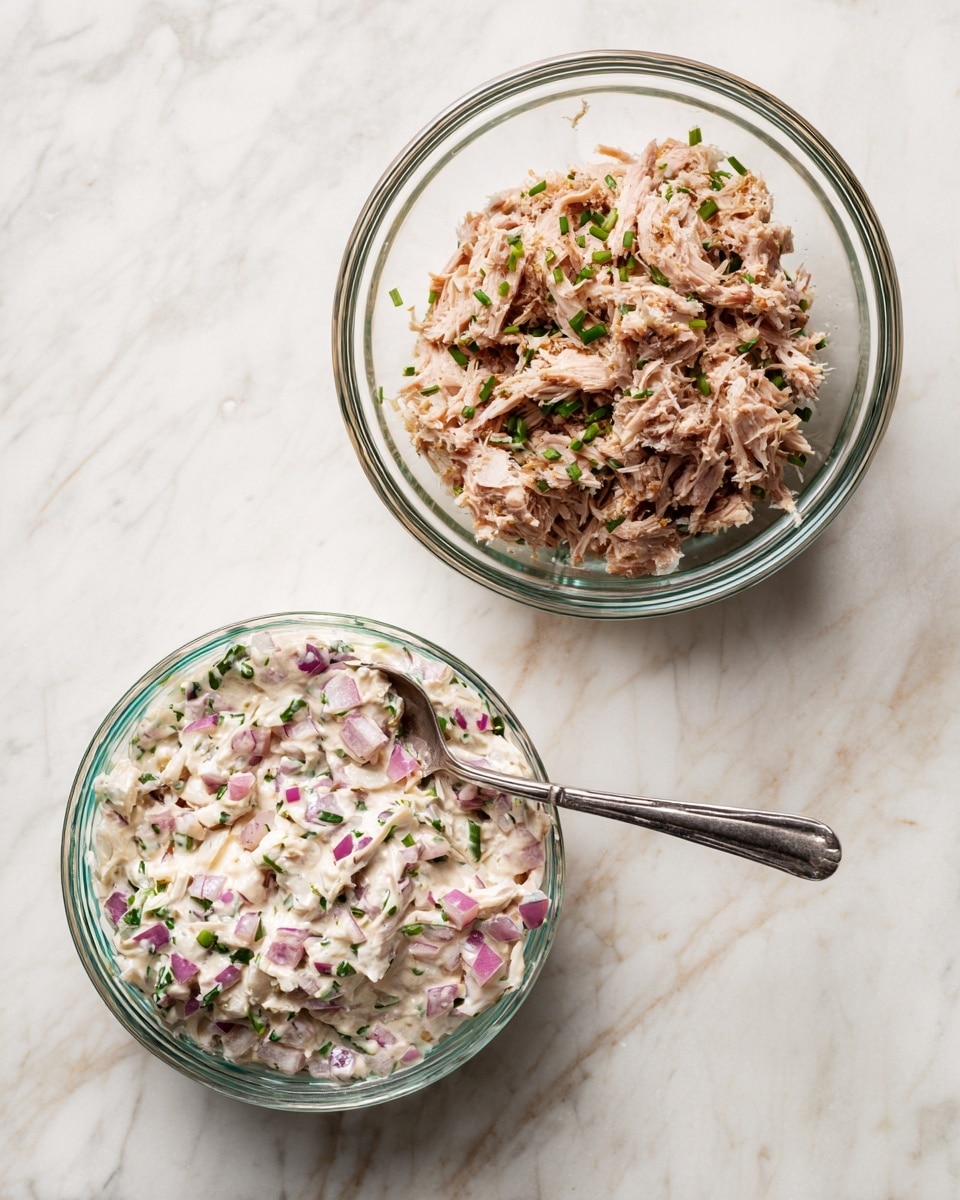 The image shows two bowls on a white marbled surface. The bowl on the left is clear glass, containing a creamy white mixture with small pieces of chopped red onions and green herbs visible throughout, and a silver spoon inside. The bowl on the right is also clear glass, filled with a mound of finely shredded meat with a mix of light brown and pale pink colors, along with some scattered small green herbs. photo taken with an iphone --ar 4:5 --v 7