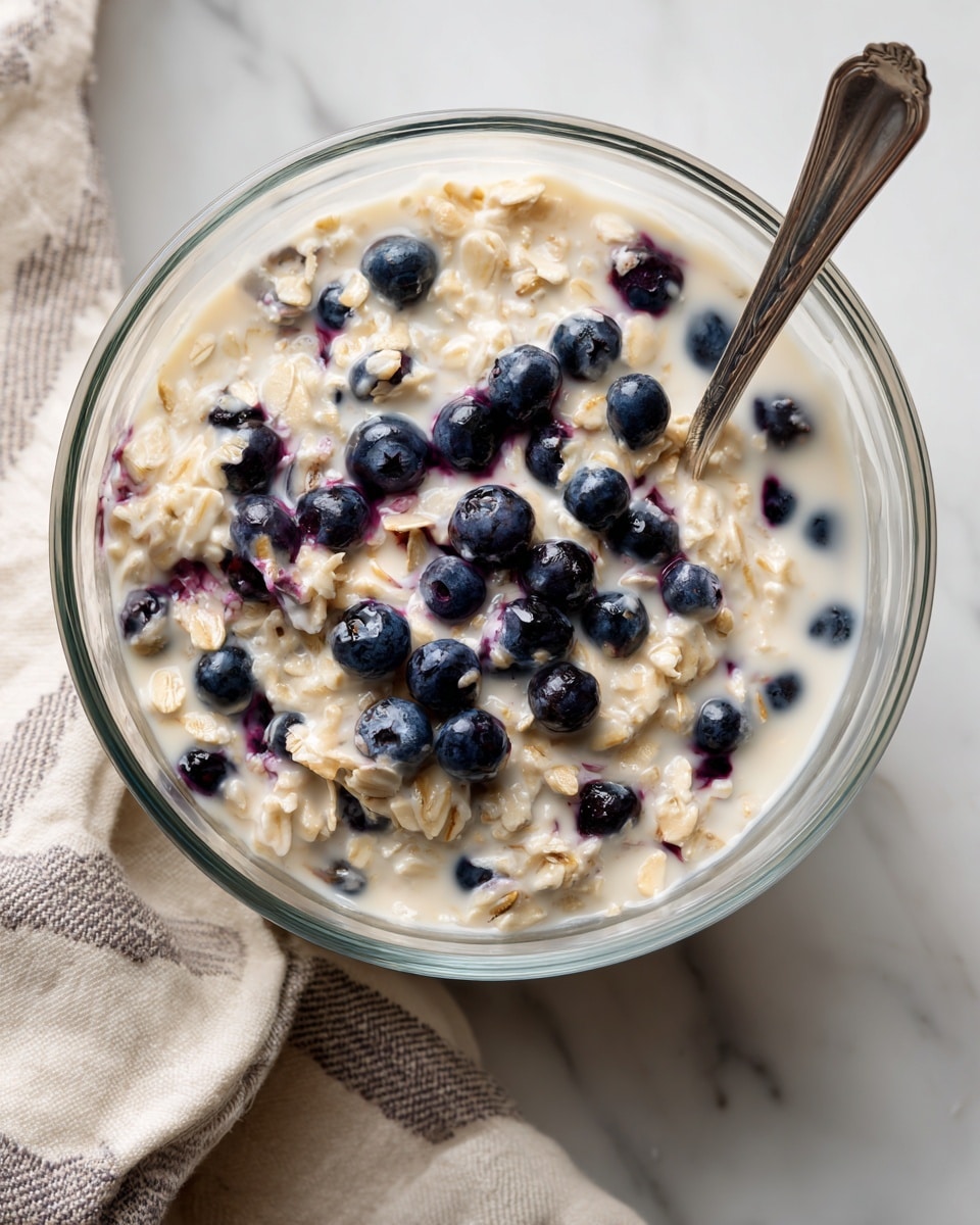 A clear glass bowl filled with a mix of creamy oats soaked in milk with scattered fresh blueberries, showing light beige and purple-blue colors, with a silver spoon resting inside the bowl partly submerged in the mixture, set on a white marbled surface with a beige and white striped cloth nearby, photo taken with an iphone --ar 4:5 --v 7