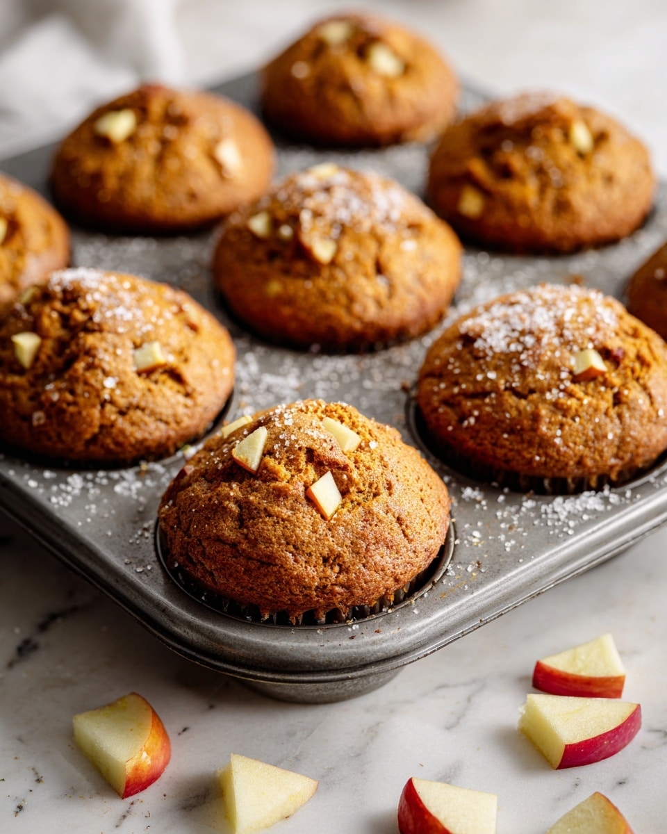 A close-up view of a metal muffin tray filled with six muffins, each golden brown in color with a slightly rough texture and small chunks of light-colored apple visible on top. The surface of the muffins is sprinkled lightly with granulated sugar, giving a sparkling effect. The tray is placed on a white marbled surface, where small pieces of diced apple and a few apple peels are scattered around. The muffins look soft and freshly baked with a smooth rounded dome shape on top. photo taken with an iphone --ar 4:5 --v 7