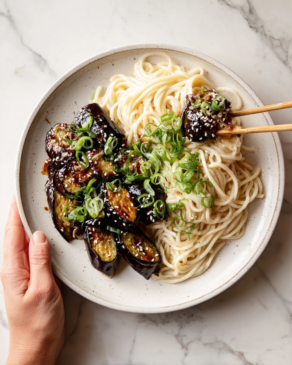 A white round plate holds three main layers: on the right side, a pile of pale creamy noodles with a soft texture; in the middle and left parts, several thick, shiny, caramel-brown roasted eggplant pieces with purple skin and tender yellow insides, topped with small white sesame seeds and bright green chopped spring onions; a pair of light wooden chopsticks held by a woman's hand are lifting one piece of eggplant, showing its glossy surface and sauce coating. The plate is on a white marbled surface. photo taken with an iphone --ar 4:5 --v 7