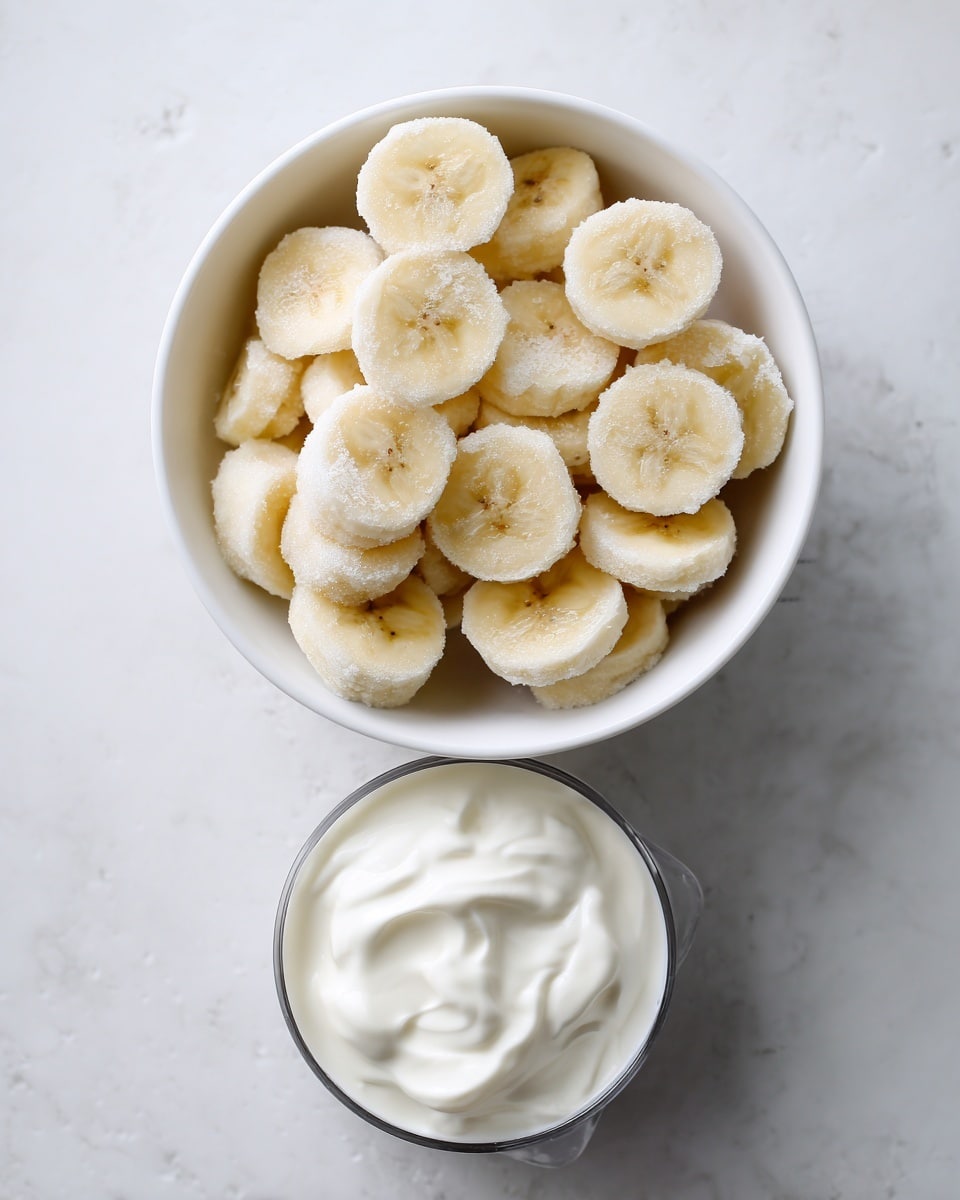 A white bowl filled with many small frozen banana slices that are pale yellow with frosty white edges, showing a rough, icy texture. Below the bowl, a metal measuring cup holds smooth, thick white plain Greek yogurt, with some textures visible on its surface. Both items are set on a white marbled textured surface. The image is simple, bright, and clean, focusing on the natural colors and textures of the frozen bananas and yogurt. Photo taken with an iphone --ar 4:5 --v 7