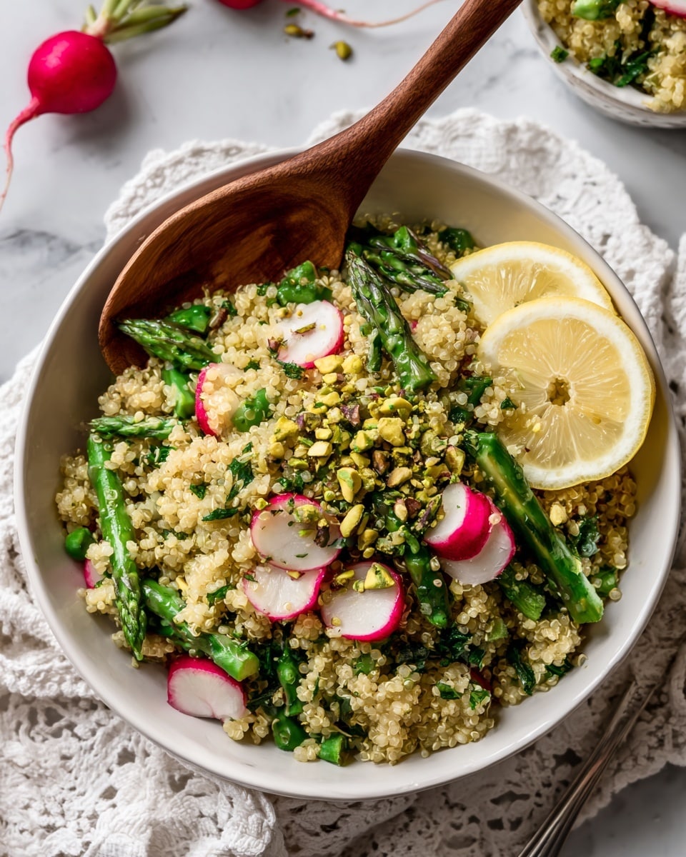 A large white bowl on a white marbled surface holds a colorful quinoa salad with visible layers of small, light beige quinoa grains mixed with green chopped vegetables like asparagus and herbs, and bright red sliced radishes throughout. The salad is topped with scattered bright green pistachio pieces and garnished with three lemon wedges placed partly on top along the edge of the bowl. To the upper right of the bowl is a clear glass filled with more chopped pistachios. A textured white cloth is placed beneath the bowl and slightly folded. Below and to the right of the bowl are two wooden salad tongs resting on the surface. The whole scene is bright and fresh, emphasizing the colors and textures of the salad. photo taken with an iphone --ar 4:5 --v 7