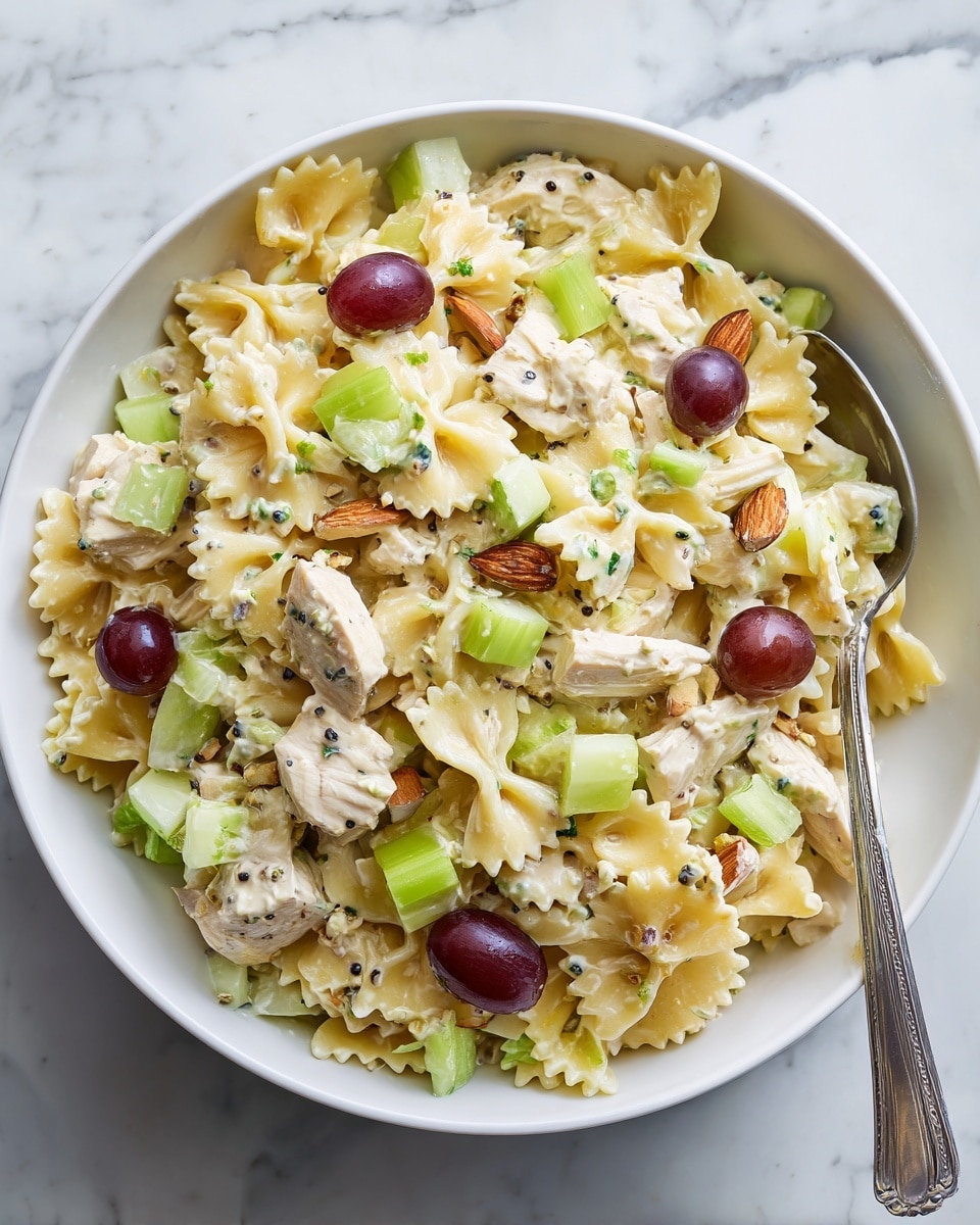 A white bowl filled with a pasta salad that has three main layers. The bottom layer is light yellow bowtie pasta mixed evenly with a creamy dressing that has small black seeds throughout. On top of the pasta, there are pieces of white chicken, chopped light green celery, and small cubes of pale green apple scattered. The highest layer has a few whole red grapes and small brown almond slices. A silver spoon is placed inside the bowl, leaning out over the edge. The bowl sits on a white marbled surface. Photo taken with an iphone --ar 4:5 --v 7