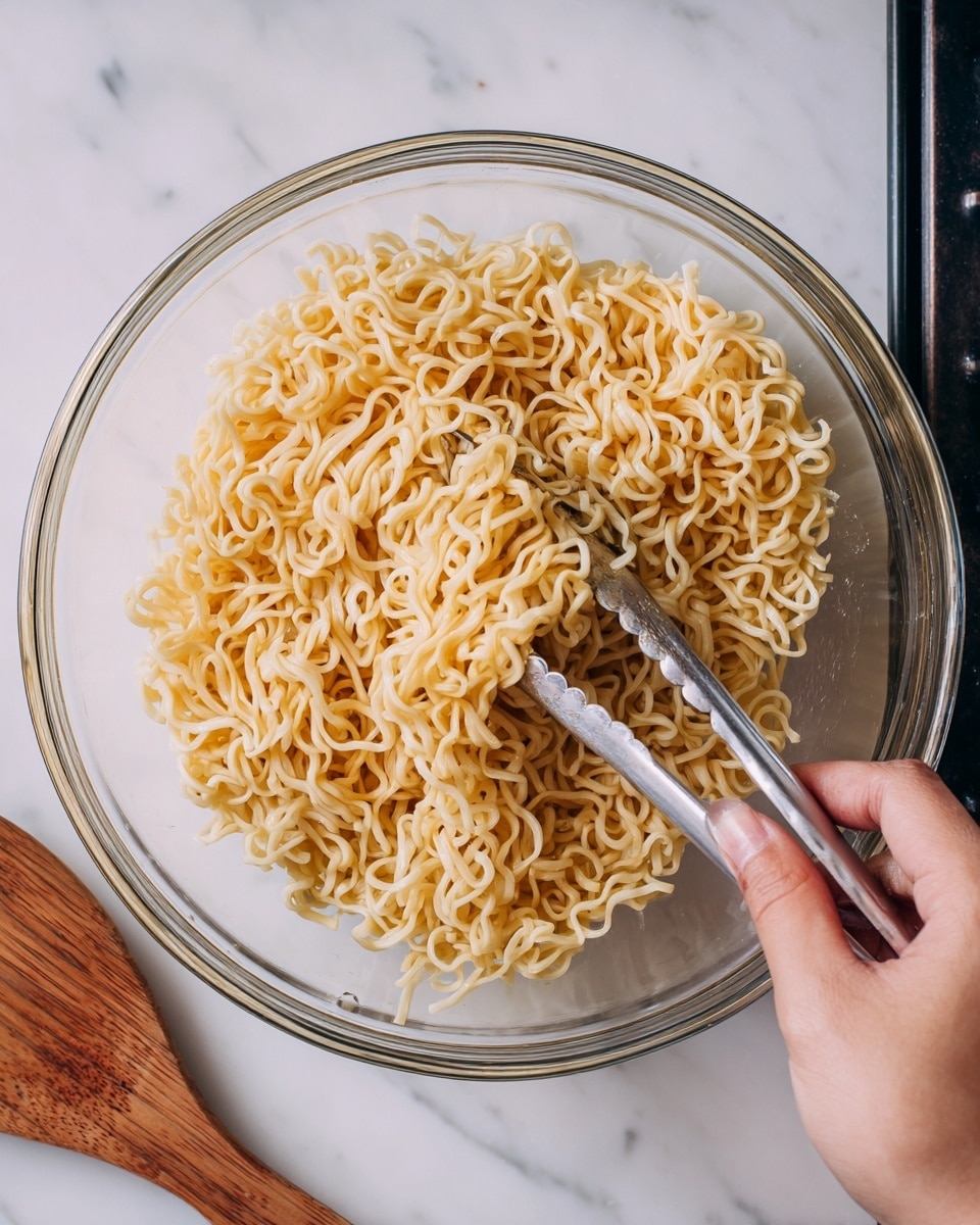 A clear glass bowl filled with light yellow instant noodles, loosely piled inside. A woman's hand is holding metal tongs, mixing the noodles from the right side of the bowl. The background shows a dark stove top and a wooden spatula resting beside the bowl. The whole scene is set on a white marbled surface. photo taken with an iphone --ar 4:5 --v 7