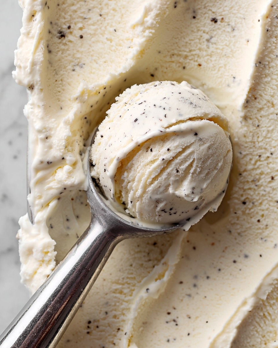 A close-up image shows a scoop of creamy, off-white vanilla ice cream with tiny black specks throughout, sitting in a shiny metal ice cream scooper. The ice cream has a smooth and slightly fluffy texture with visible small air pockets and some uneven areas where the scooper has moved through it. The background features a broad flat layer of the same vanilla ice cream spread evenly across a surface with a soft, smooth texture and specks scattered in it. The setting has a clean white marbled texture beneath the ice cream. photo taken with an iphone --ar 4:5 --v 7