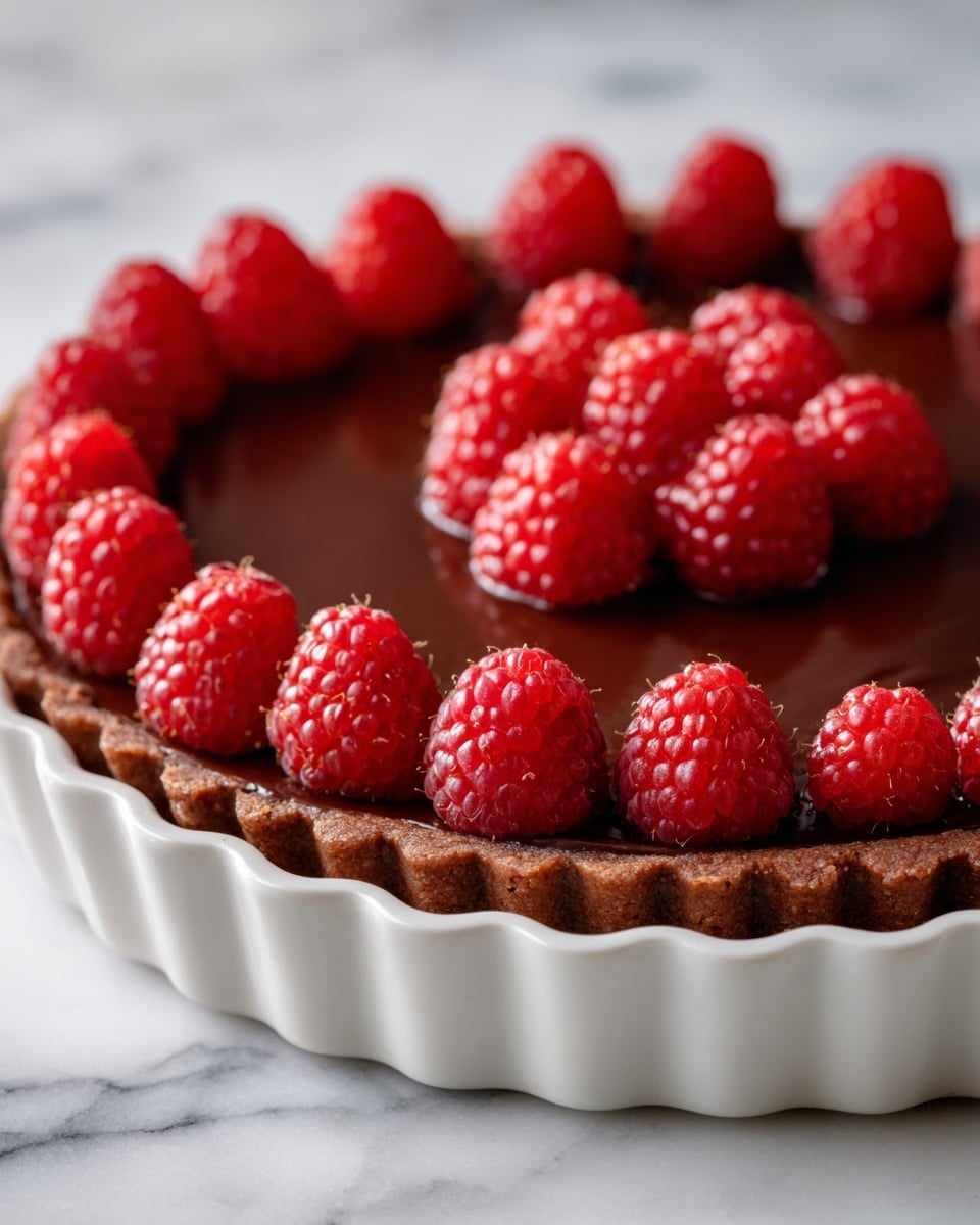 The dessert has four clear layers, starting from the bottom with a dark crumbly crust, followed by a thick dark brown creamy layer, then a lighter beige creamy layer, and topped with a shiny dark chocolate ganache. On the top edge, there is a row of bright red raspberries evenly spaced around the cake. The cake is on a white fluted cake stand with a white marbled surface in the background. Photo taken with an iphone --ar 4:5 --v 7