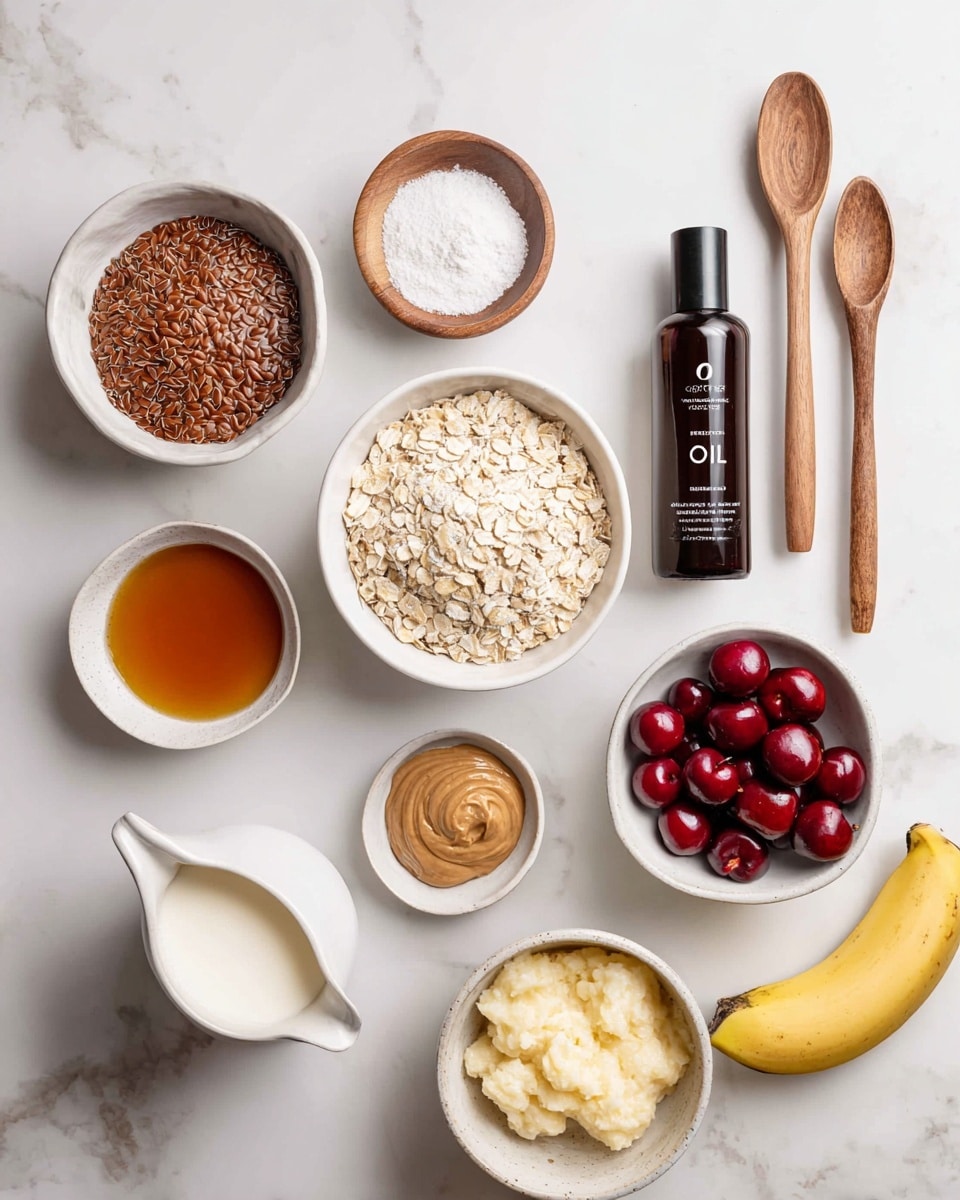 The image shows an arrangement of various small white bowls and containers on a white marbled surface. From left to right on the top row, there is a white bowl filled with brown flaxseeds, two wooden spoons standing vertically with a small amount of vanilla and almond extract in them, a small round wooden bowl containing white baking powder, and a white bowl filled with amber-colored maple syrup. Below them, there is a white bowl filled with light brown nut butter and a small white round bowl with frozen bright red cherries. A large white bowl filled with pale golden oats sits centrally, with a small dark bottle labeled