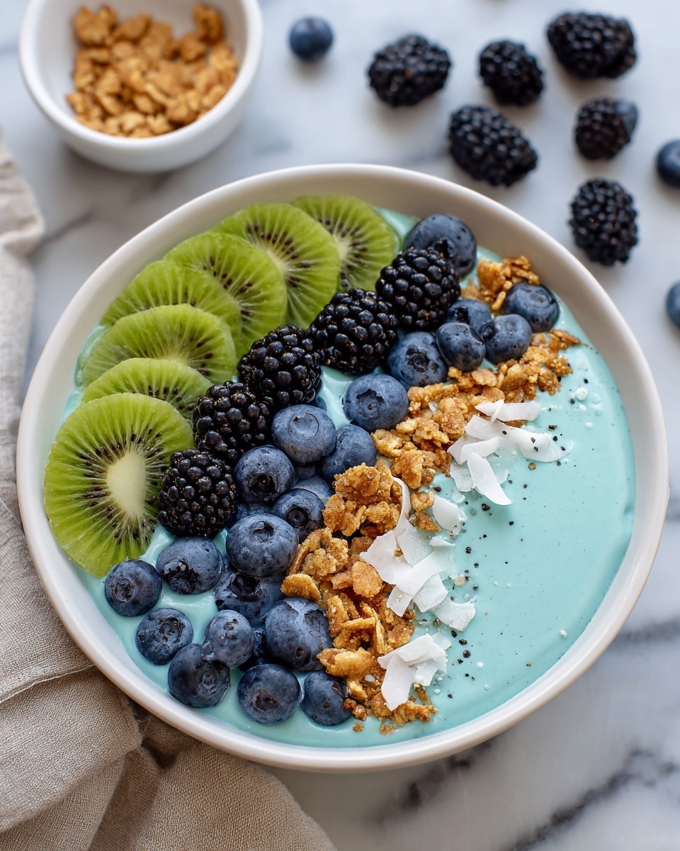 A white bowl filled with a bright blue creamy base takes up most of the image. On top, there are several thin slices of green kiwi arranged neatly on the left edge. Next to the kiwi are clusters of golden brown granola pieces. Around the granola are plump, dark blackberries and round blueberries, adding deep purple and blue colors to the bowl. There are also some white coconut flakes scattered near the blackberries. The bowl rests on a white marbled surface, and some loose blackberries and blueberries are scattered in the background. The scene includes a corner of a small white bowl with more coconut flakes and a beige cloth at the front edge. photo taken with an iphone --ar 4:5 --v 7