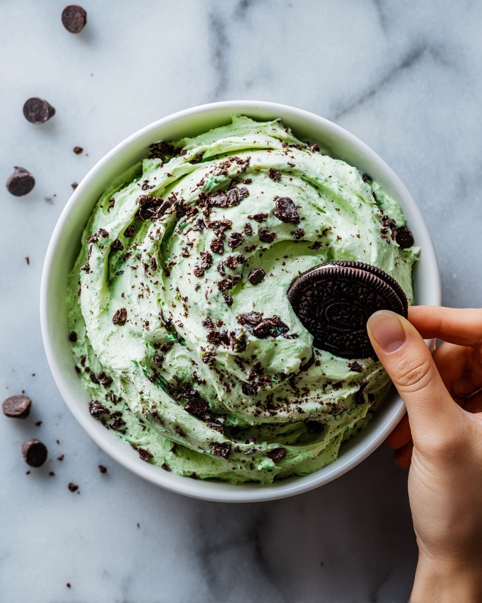 A close-up of a woman's hand holding a chocolate Oreo cookie with a generous scoop of light green mint cream mixed with small dark chocolate chips on top. The background shows a white bowl filled with the same mint cream mixture that has scattered small dark chocolate chips on its light green fluffy surface. The whole scene is set on a white marbled background, bright and clean. photo taken with an iphone --ar 4:5 --v 7