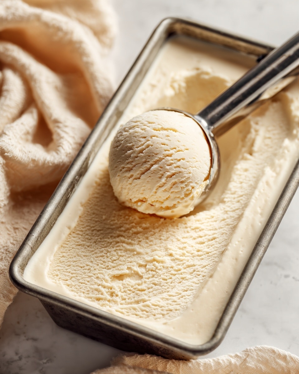 A close-up image of a metal rectangular container filled with smooth, creamy light beige ice cream. A shiny metal ice cream scooper holds a single rounded scoop of the ice cream above the container, showing its soft, slightly textured surface. The background has a soft, beige cloth gently wrinkled on a white marbled surface, adding softness to the setting. photo taken with an iphone --ar 4:5 --v 7