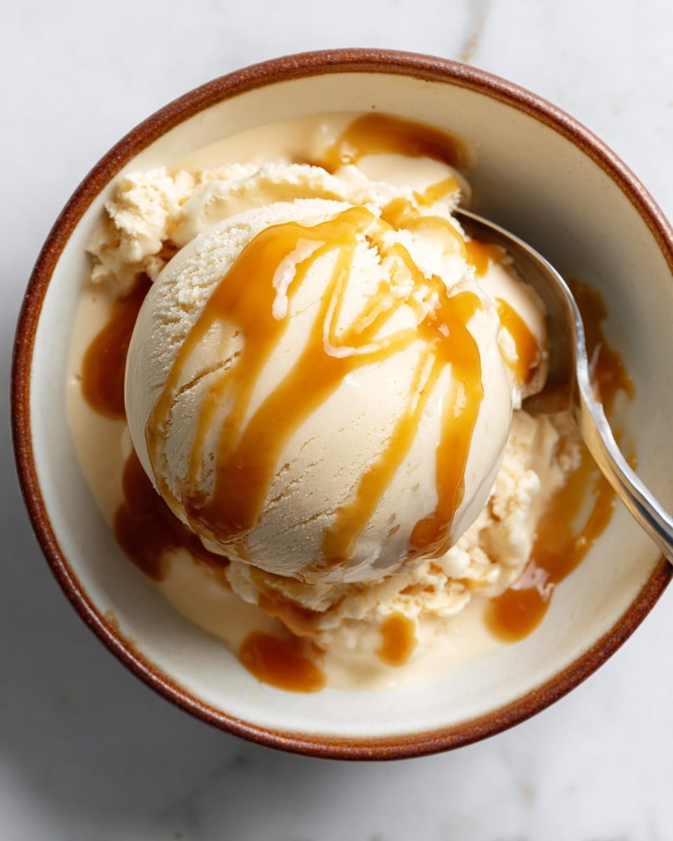 A white bowl with a brown rim holds a smooth scoop of creamy white dessert that has a few shiny, orange patches of sauce drizzled on top. A silver spoon is scooping into the dessert, showing its soft texture. Next to the bowl, there is a small clear glass container holding a thick orange sauce, with some sauce clinging to the edges, and beside it, a wooden lid with a natural bamboo pattern rests on a white marbled surface. Photo taken with an iphone --ar 4:5 --v 7