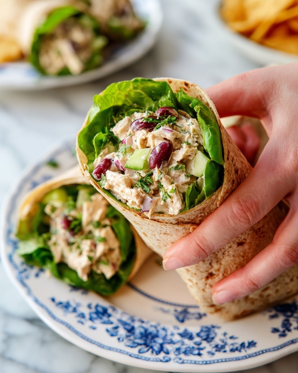 A close-up of a wrap held by a woman's hand showing three main layers: a soft, lightly toasted brownish tortilla on the outside, a middle layer of bright green lettuce leaves, and a creamy, chunky filling made of beige chicken salad mixed with small pieces of red kidney beans, green celery, bits of red onion, and fresh green herbs sprinkled on top. The wrap is on a white plate with a blue floral pattern resting on a white marbled surface, with more wraps filled with the same ingredients arranged around it and some tortilla chips placed in the background. Photo taken with an iphone --ar 4:5 --v 7