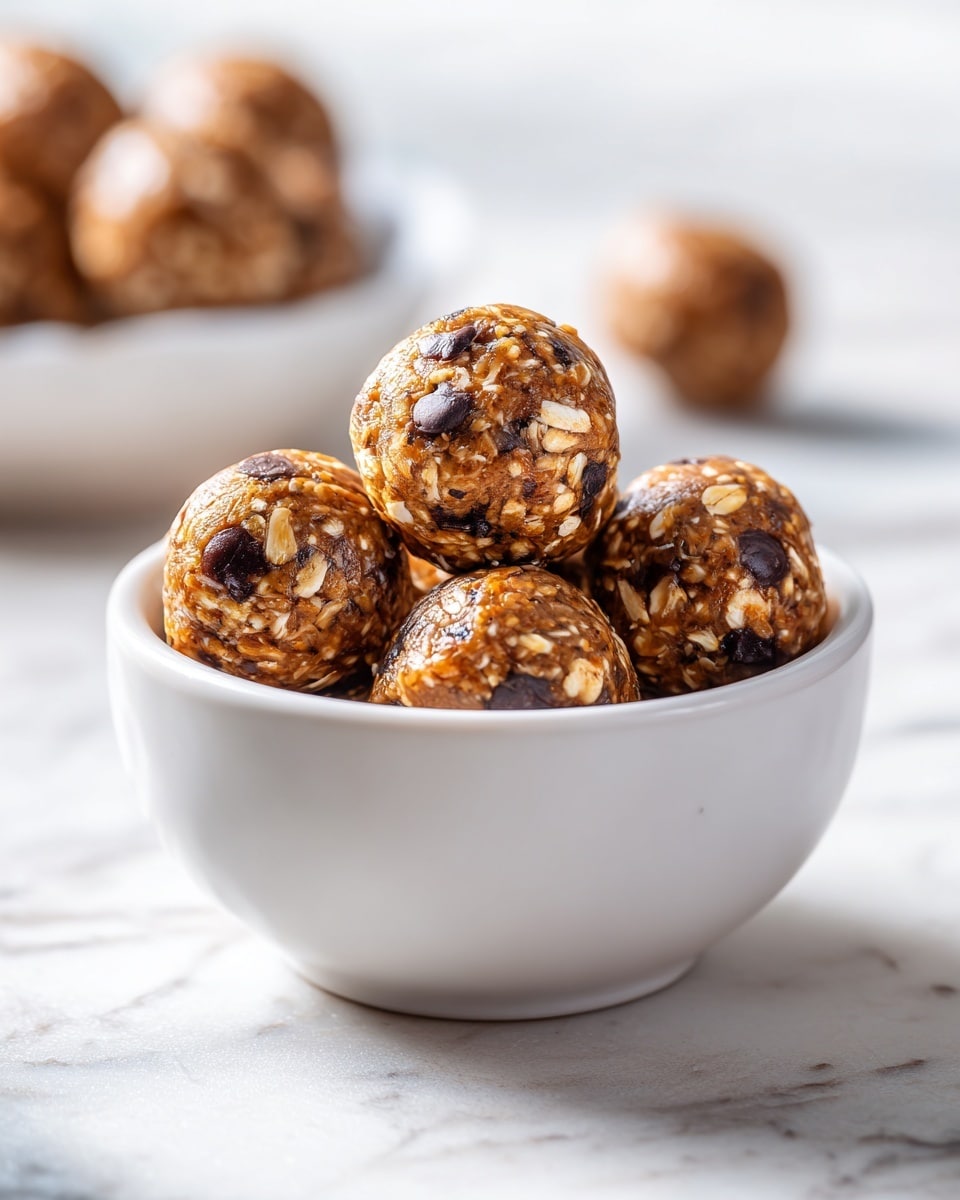 A white bowl is filled with round, golden-brown energy balls dotted with small, dark chocolate chips and bits of oats, showing a rough, textured surface. One energy ball is placed on top, centered in the bowl, revealing a clear view of its oat and chocolate chip-filled interior. The bowl sits on a white marble textured surface, and a blurred white bowl with more energy balls is visible in the top left corner. A single energy ball is also out of focus in the bottom right corner. The lighting is bright and natural, highlighting the warm colors and textures of the energy balls. Photo taken with an iphone --ar 4:5 --v 7