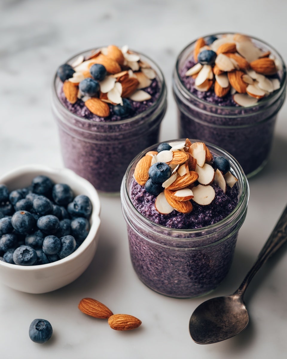 The image shows three clear glass jars filled with a thick, purple oatmeal-like mixture. Each jar is topped with a layer of fresh, plump blueberries and scattered slices of toasted almonds. Around the jars are two white bowls, one filled with whole blueberries and the other with almond slices. There is a dark metallic spoon near the bowls. The whole scene is set on a white marbled surface, giving a clean and fresh look. photo taken with an iphone --ar 4:5 --v 7