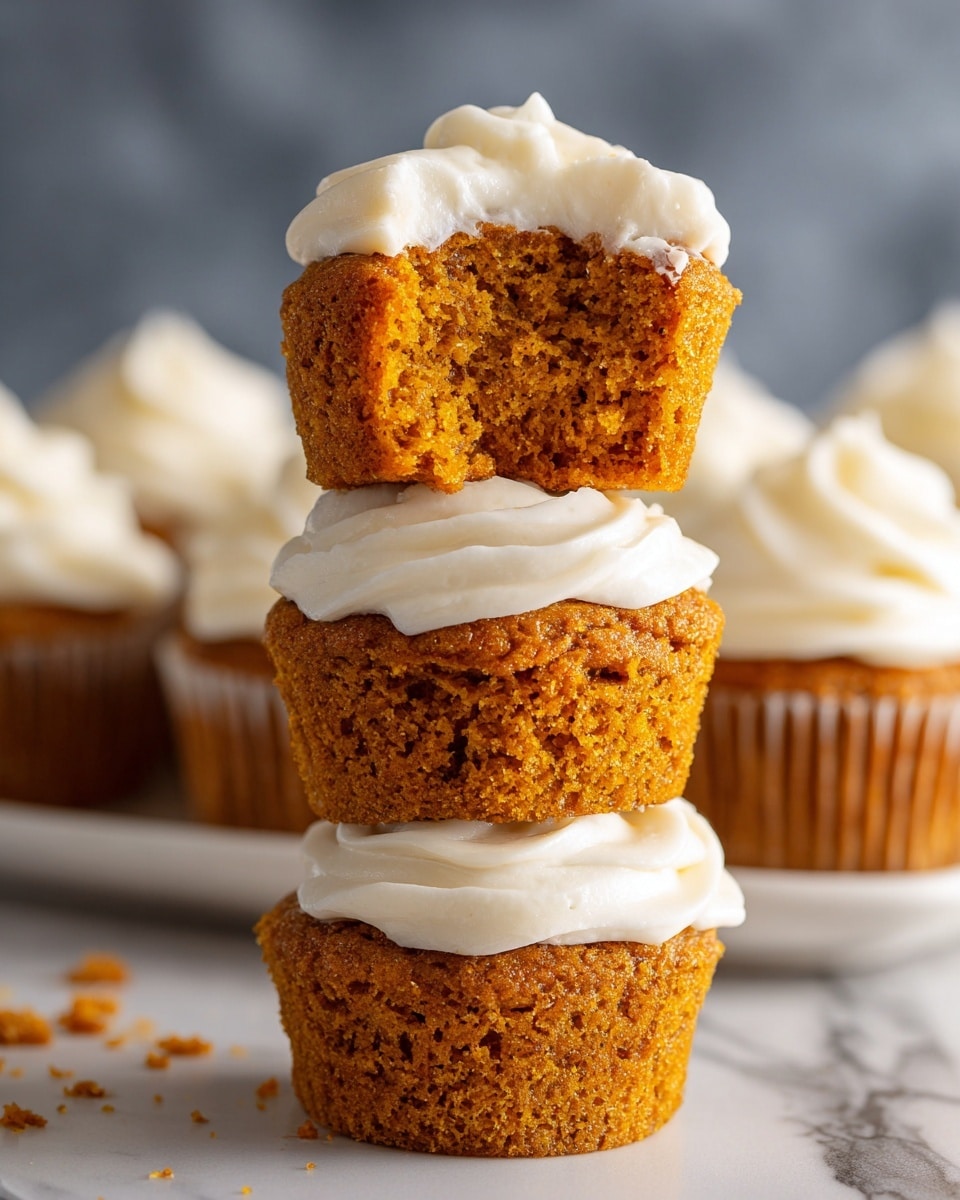 The image shows a close-up of three stacked pumpkin muffins with a thick layer of white frosting on top of each one. The muffins are bright orange with a moist, crumbly texture. The top muffin has a bite taken out of it, revealing its soft inside. Below the stack, there is a white tray filled with many pumpkin muffins, each topped with smooth, swirled white frosting. The background has a soft, dark blur with a white marbled surface under the muffins. photo taken with an iphone --ar 4:5 --v 7