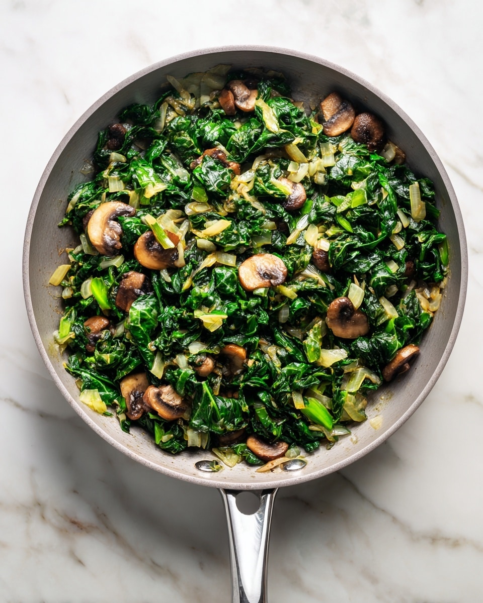 A deep pan filled with a cooked mix of chopped dark green leafy vegetables and small light brown mushroom slices, combined with translucent pale yellow onion pieces throughout. The greens and mushrooms create a textured, uneven but well-mixed layer that covers the bottom of the pan. The pan itself is light gray with a shiny metal handle and is set on a white marbled surface. Photo taken with an iphone --ar 4:5 --v 7