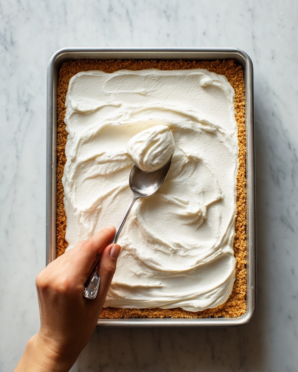 A close-up view of a rectangular dessert with two layers: a bottom layer of golden-brown crust with a crumbly texture, and a top layer of smooth, white creamy spread being evenly smoothed out by a woman's hand holding a silver spoon. The dessert is in a metal baking tray placed on a white marbled surface. The creamy layer shows soft swirls and peaks where it is being spread. photo taken with an iphone --ar 4:5 --v 7